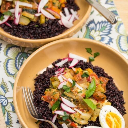 Overhead view of black rice bowls topped with veggies, herbs, and eggs.