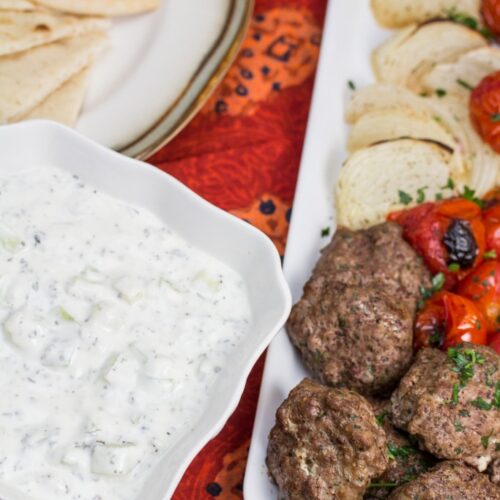 Overhead view of grilled meat, veggies, pita bread, and a bowl of mast-o-khiar yogurt sauce.