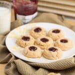 Thumbprint cookies on a plate with a glass of milk and jar of jelly in the background.