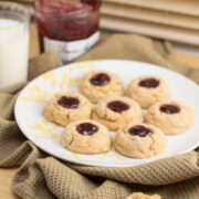 Thumbprint cookies on a plate with a glass of milk and jar of jelly in the background.