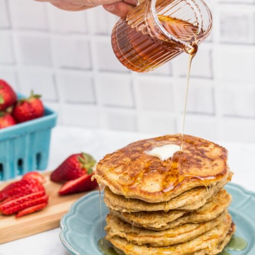 Blue plate holding a stack of buttermilk pancakes with syrup being poured over top and strawberries in the background.