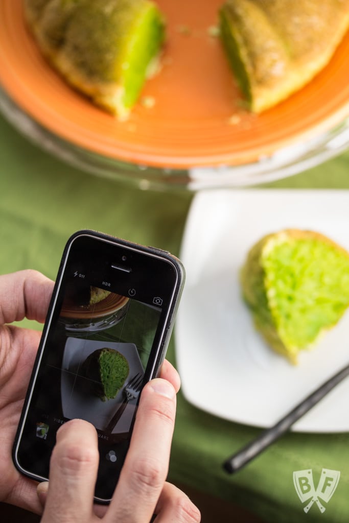 Hands taking a photo with a phone of a green bundt cake on a plate with a slice placed on a smaller plate with a vibrant green filling.