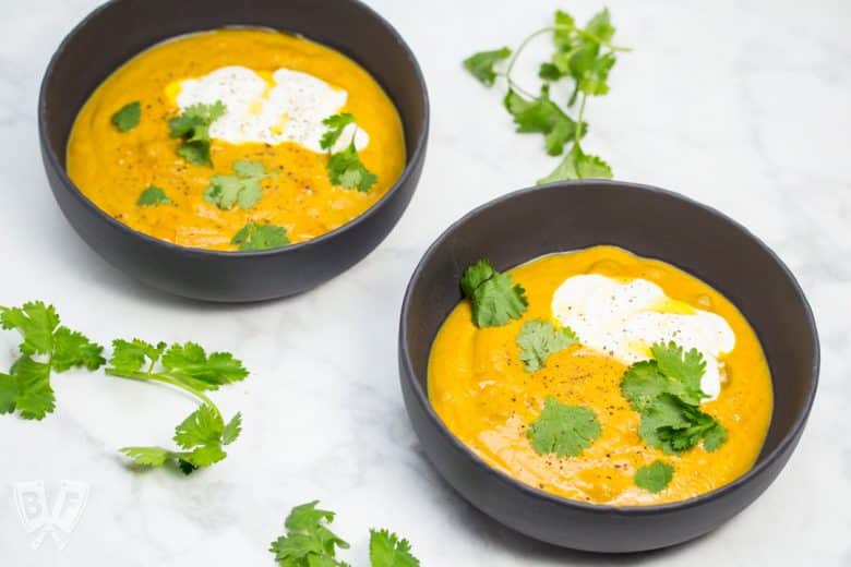 Overhead view of 2 bowls of curried pumpkin soup with cilantro leaves, sour cream, and chili oil garnish.