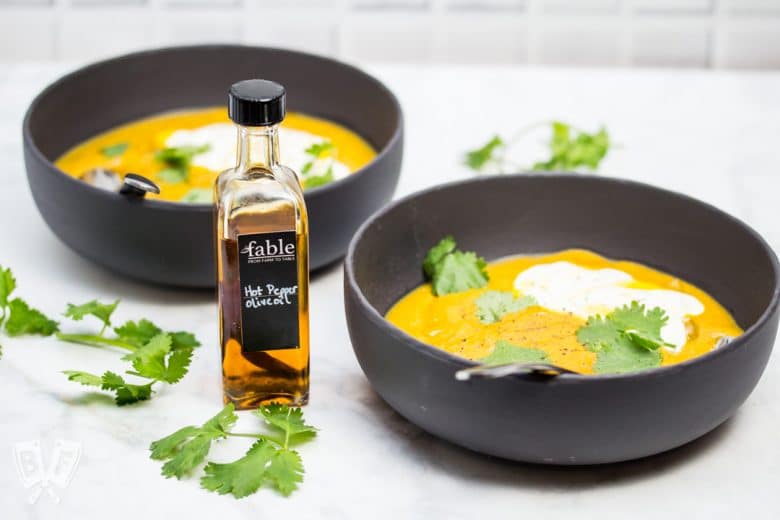 ¾ view of 2 bowls of curried pumpkin soup with cilantro leaves, sour cream, and a bottle of chili oil.