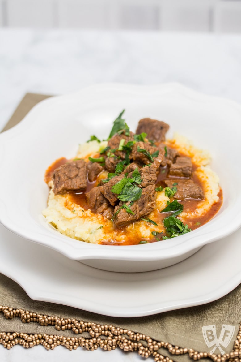 A heaping portion of mashed potatoes in a bowl with beef goulash poured over top, sprinkled with parsley.