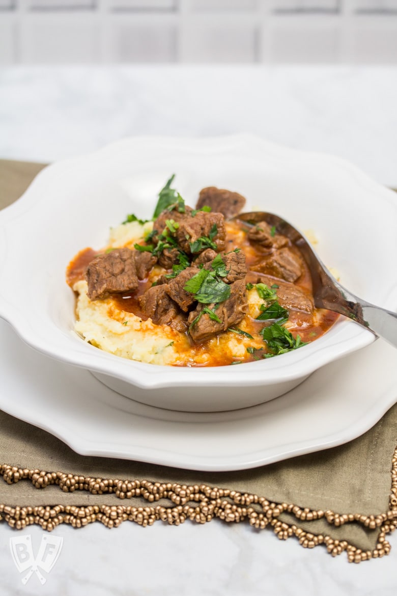 A heaping portion of mashed potatoes in a bowl with beef goulash poured over top, sprinkled with parsley.