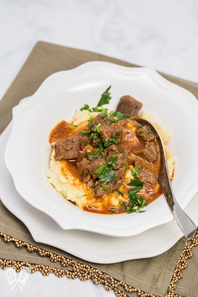 A heaping portion of mashed potatoes in a bowl with beef goulash poured over top, sprinkled with parsley.