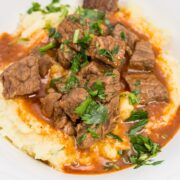 Close-up view of a heaping portion of mashed potatoes in a bowl with beef goulash poured over top, sprinkled with parsley.