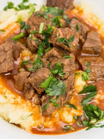 Close-up view of a heaping portion of mashed potatoes in a bowl with beef goulash poured over top, sprinkled with parsley.