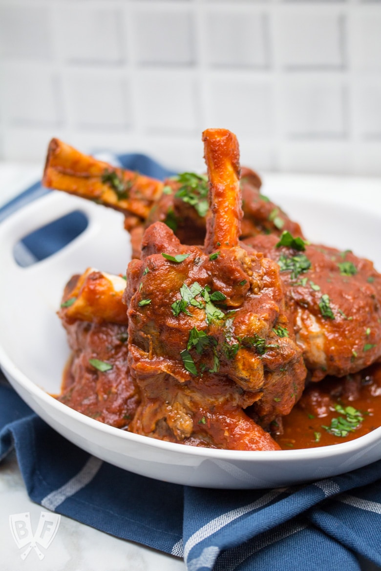 ¾ view of lamb shanks with tomato sauce sprinkled with parsley in a serving bowl.