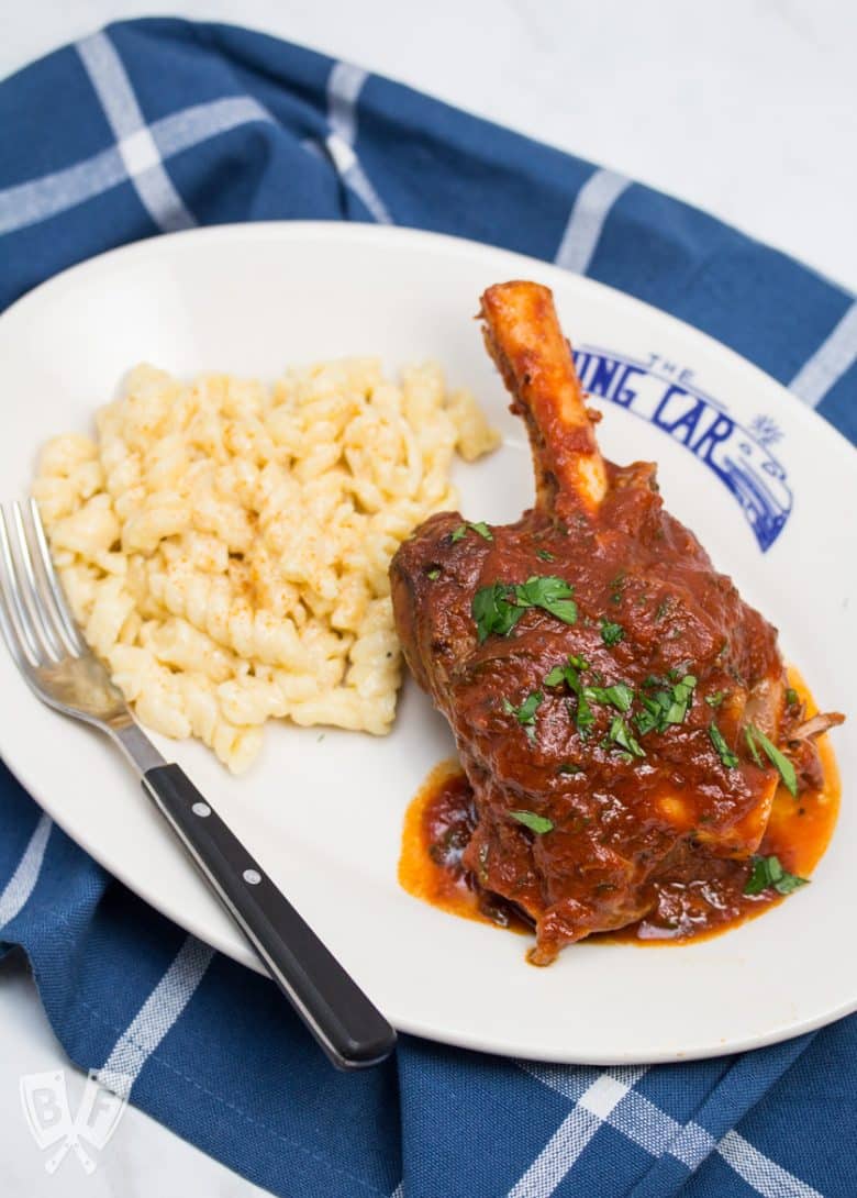 Overhead view of a plate with a lamb shank and tomato sauce next to macaroni and cheese with a fork on the plate.