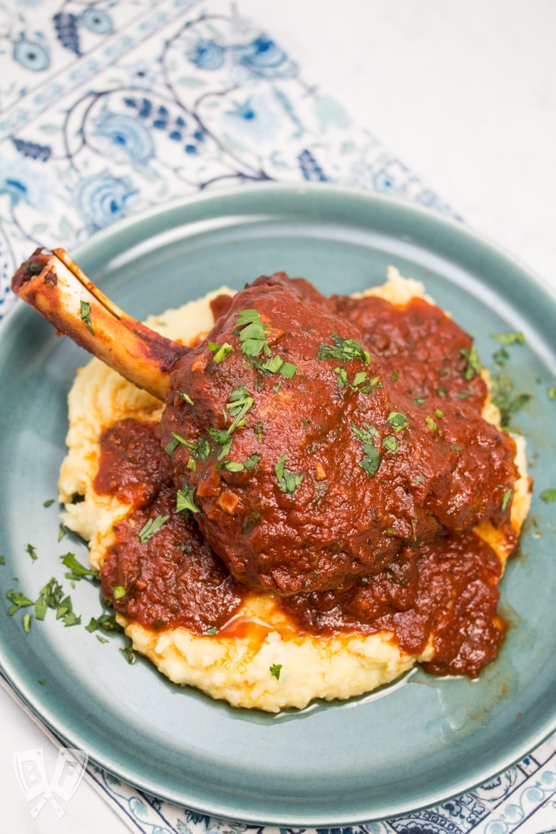 Overhead view of a plate with mashed potatoes topped with a lamb shank with tomato sauce sprinkled with parsley.