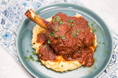 Overhead view of a plate with mashed potatoes topped with a lamb shank with tomato sauce sprinkled with parsley.