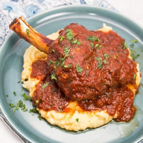 Overhead view of a plate with mashed potatoes topped with a lamb shank with tomato sauce sprinkled with parsley.