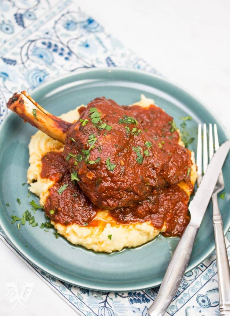 Overhead view of a plate with mashed potatoes topped with a lamb shank with tomato sauce sprinkled with parsley and fork and knife off to the side.