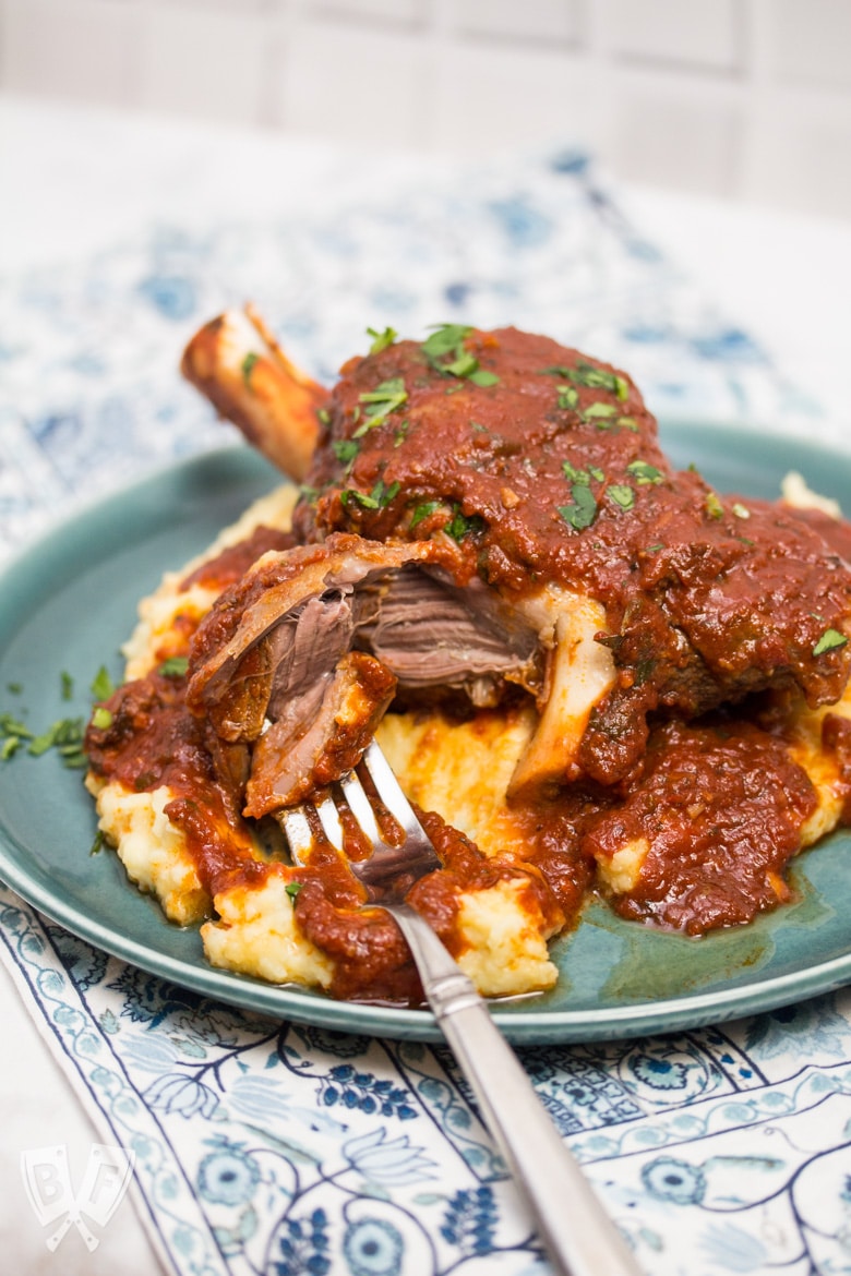 3/4 view of a plate with mashed potatoes topped with a lamb shank with tomato sauce sprinkled with parsley. A fork is pulling the meat off the bone.