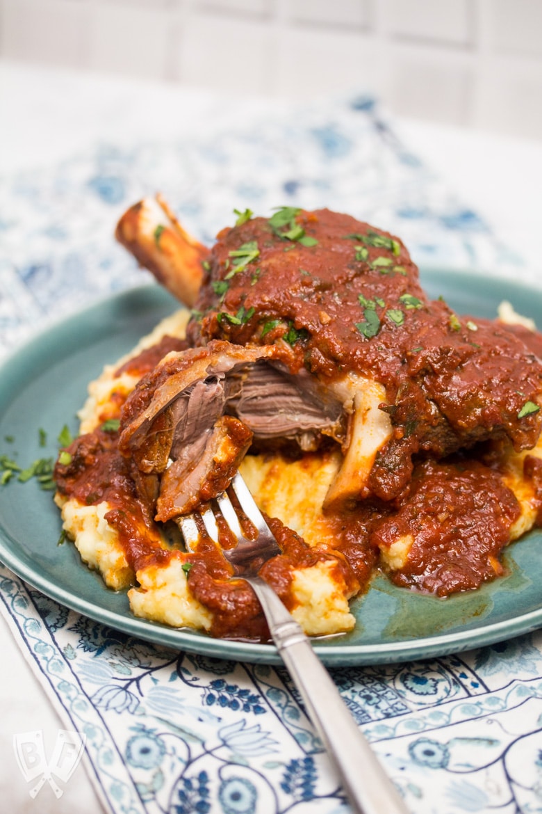 ¾ view of a plate with mashed potatoes topped with a lamb shank with tomato sauce sprinkled with parsley. A fork is pulling the meat off the bone.