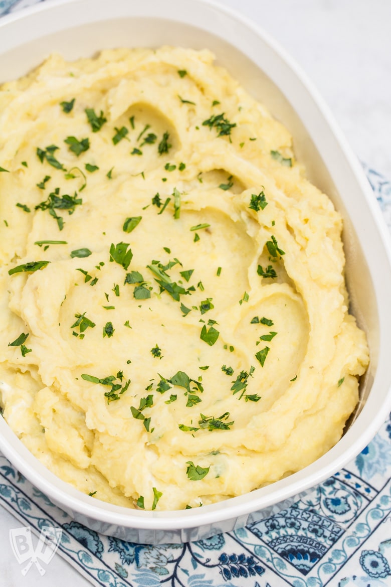 Overhead shot of an oval casserole dish full of creamy mashed potatoes sprinkled with chopped parsley.