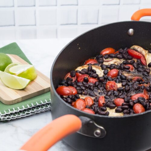 ¾ view up of a Latin Chicken Skillet with Black Beans + Tomatoes with a cutting board and limes off to the side.