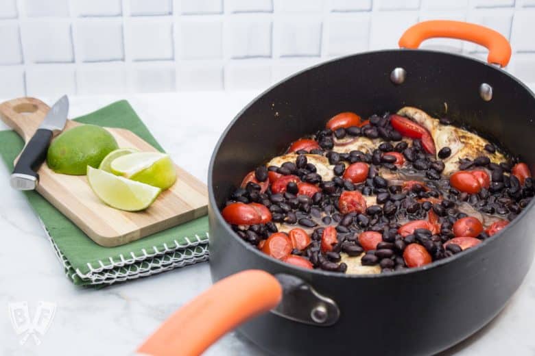 ¾ view up of a Latin Chicken Skillet with Black Beans + Tomatoes with a cutting board and limes off to the side.