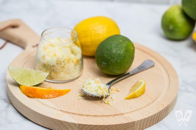 A round cutting board with fresh citrus and a spoon and jar full of a citrus zest, salt, and sugar mixture.