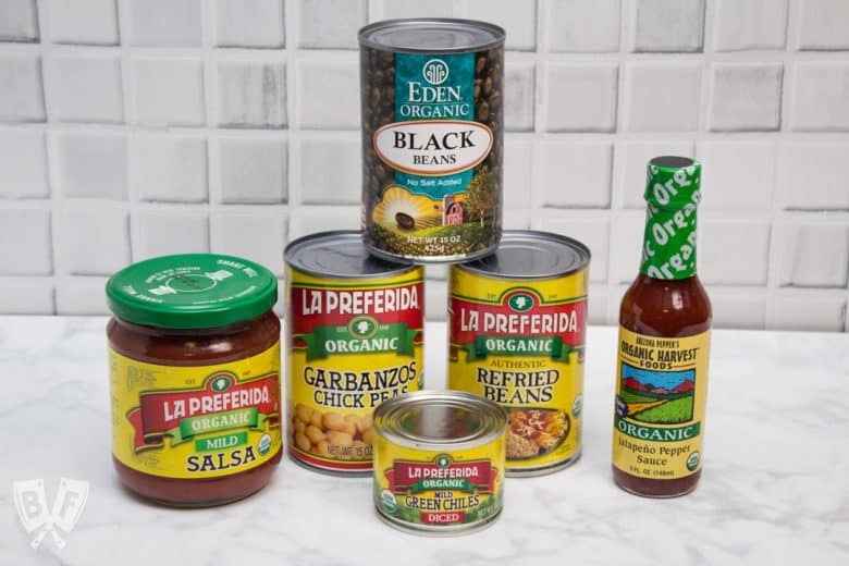 A stack of canned beans, salsa, hot sauce, and chiles displayed on a kitchen counter.