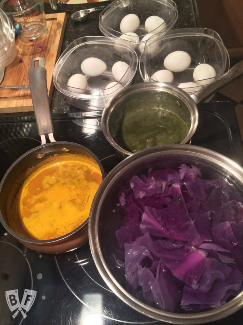 Overhead view of pots full of natural Easter egg dye ingredients with bowls of hard boiled eggs in the background.