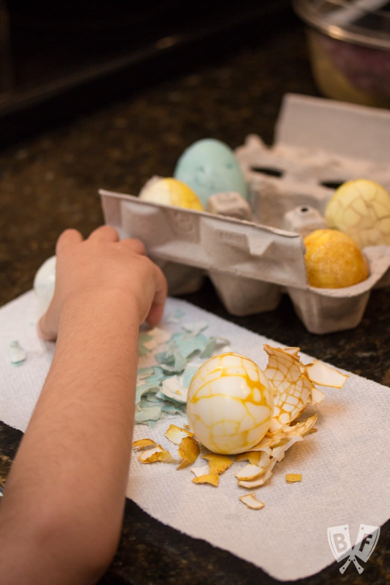 A child's hand holding a naturally dyed Easter egg with a cracked shell pattern on it and more Easter eggs in a carton in the background.