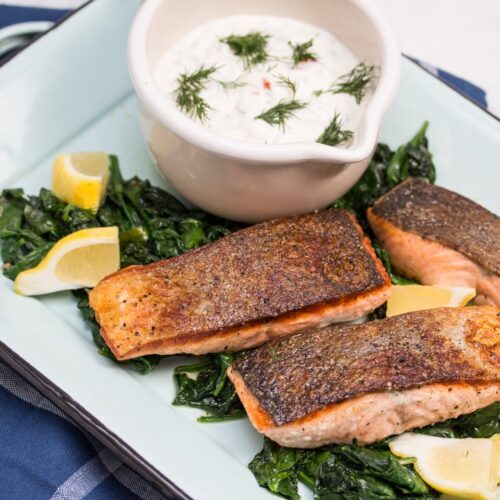 Overhead view of a platter of seared salmon on top of spinach and lemon wedges with a bowl of sauce in the background.