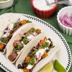 Overhead view of prepared ingredients and 3 assembled Roasted Sweet Potato and Black Bean Tacos with Cilantro Chimichurri.