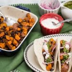 Overhead view of prepared ingredients and 3 assembled Roasted Sweet Potato and Black Bean Tacos with Cilantro Chimichurri.