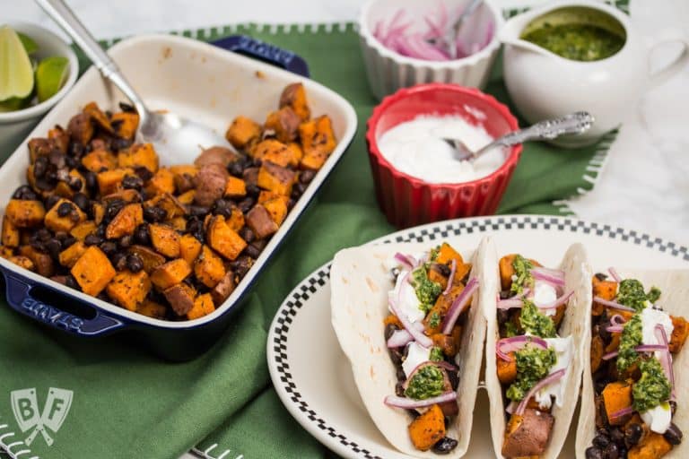 Overhead view of prepared ingredients and 3 assembled Roasted Sweet Potato and Black Bean Tacos with Cilantro Chimichurri.