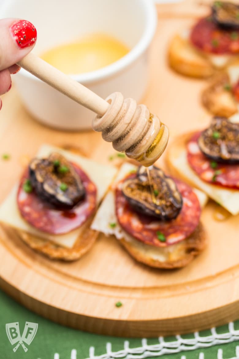 A hand holding a honey dipper drizzling honey onto Chorizo & Fig Crostini displayed on a board.