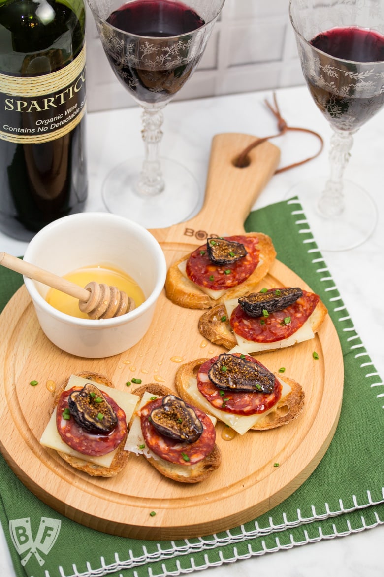 Honeyed Chorizo & Fig Crostini displayed on a board with a honey dipper and a bottle & 2 glasses of Spanish wine.