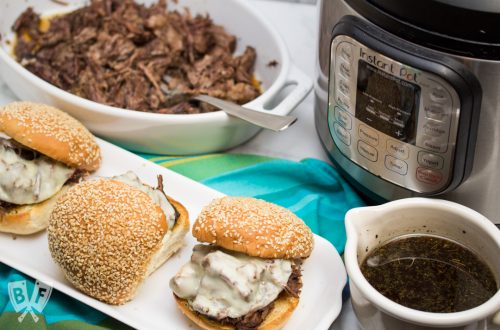 3/4 view of 3 assembled Instant Pot French Dip Sandwiches with au jus, an Instant Pot, and shredded meat alongside.