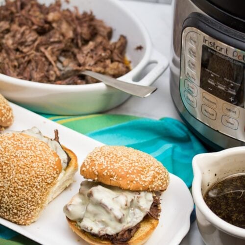 ¾ view of 3 assembled Instant Pot French Dip Sandwiches with au jus, an Instant Pot, and shredded meat alongside.