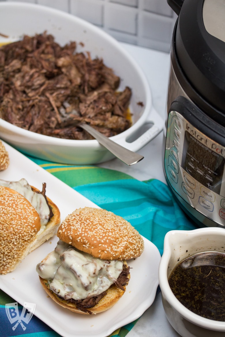 ¾ view of 3 assembled Instant Pot French Dip Sandwiches with au jus, an Instant Pot, and shredded meat alongside.