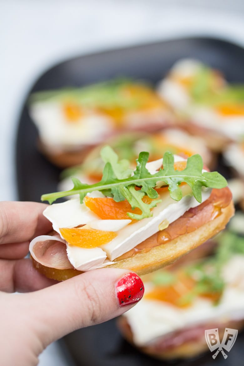 Close-up view of a hand holding a Prosciutto & Brie Crostini with Dried Apricots and a platter in the background.