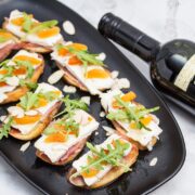 Overhead view of a platter of Prosciutto & Brie Crostini with Dried Apricots next to a bottle of red wine.