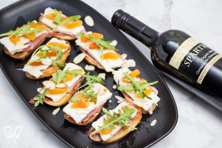 Overhead view of a platter of Prosciutto & Brie Crostini with Dried Apricots next to a bottle of red wine.