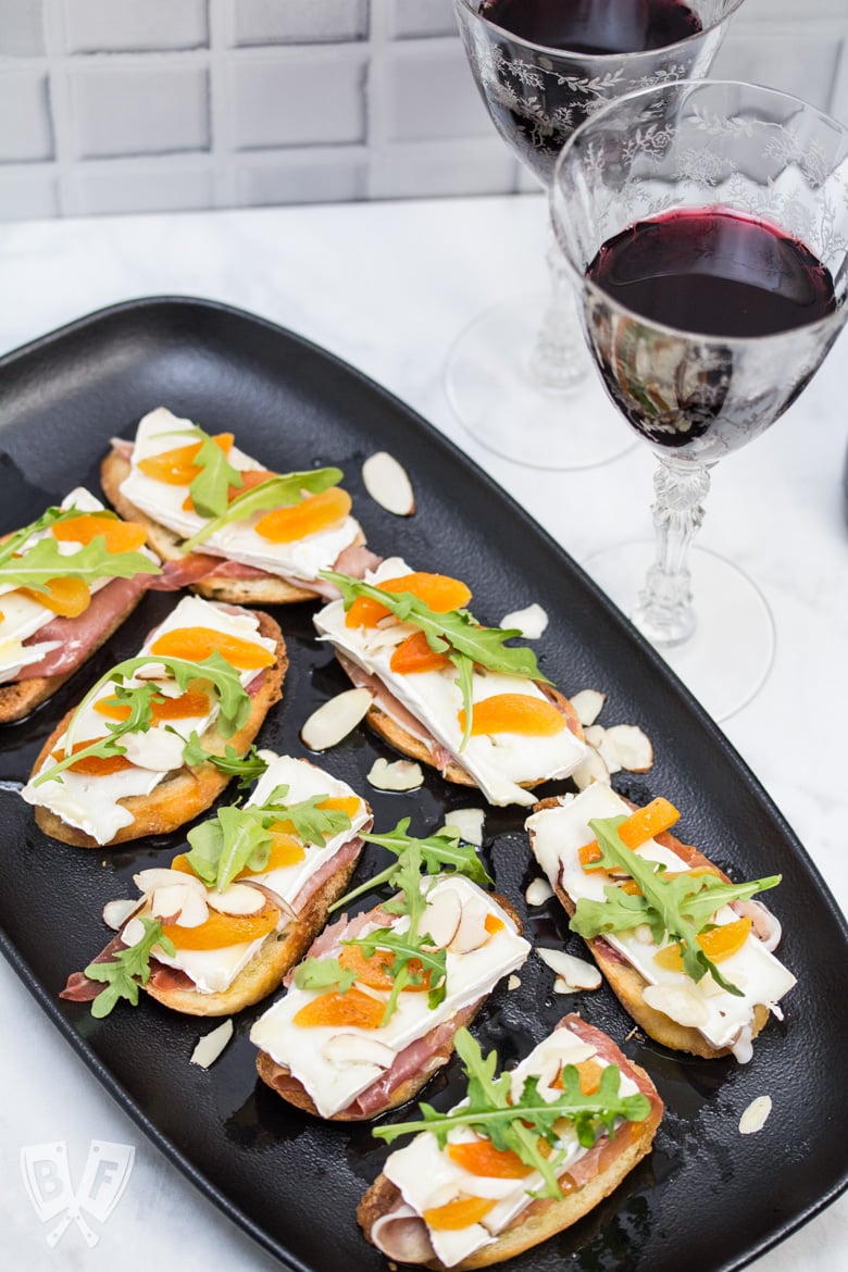 Overhead view of a platter of Prosciutto & Brie Crostini with Dried Apricots next to 2 glasses of red wine.