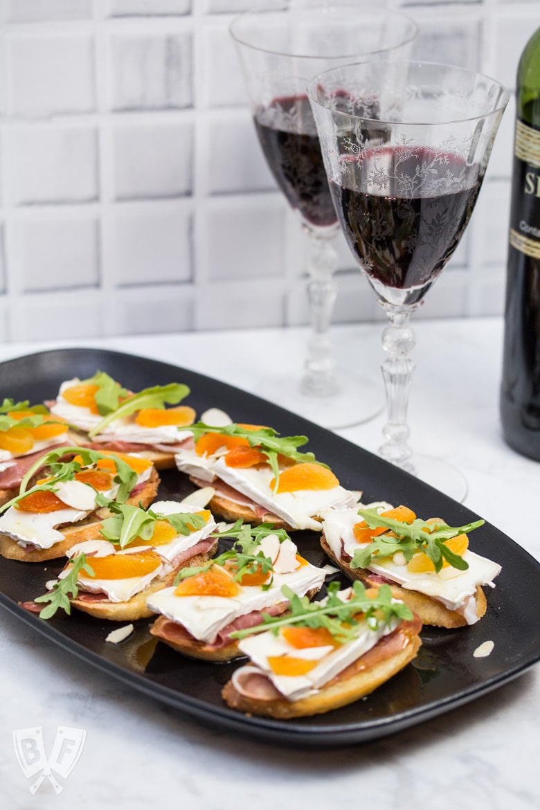 ¾ view of a platter of Prosciutto & Brie Crostini with Dried Apricots next to a bottle of red wine.