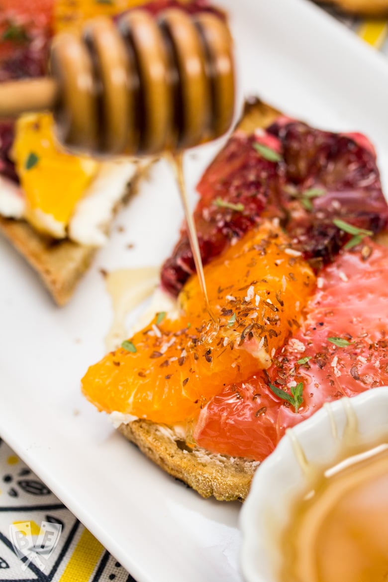 Honey dipper drizzling honey onto Goat Cheese Toast with Honeyed Citrus Fruit