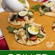 Pouring vinaigrette over a tray of mini naan breads topped with grilled Niçoise salads.