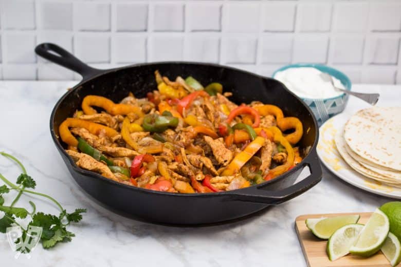 A cast iron skillet filled with chicken fajitas with tortillas and garnishes alongside.