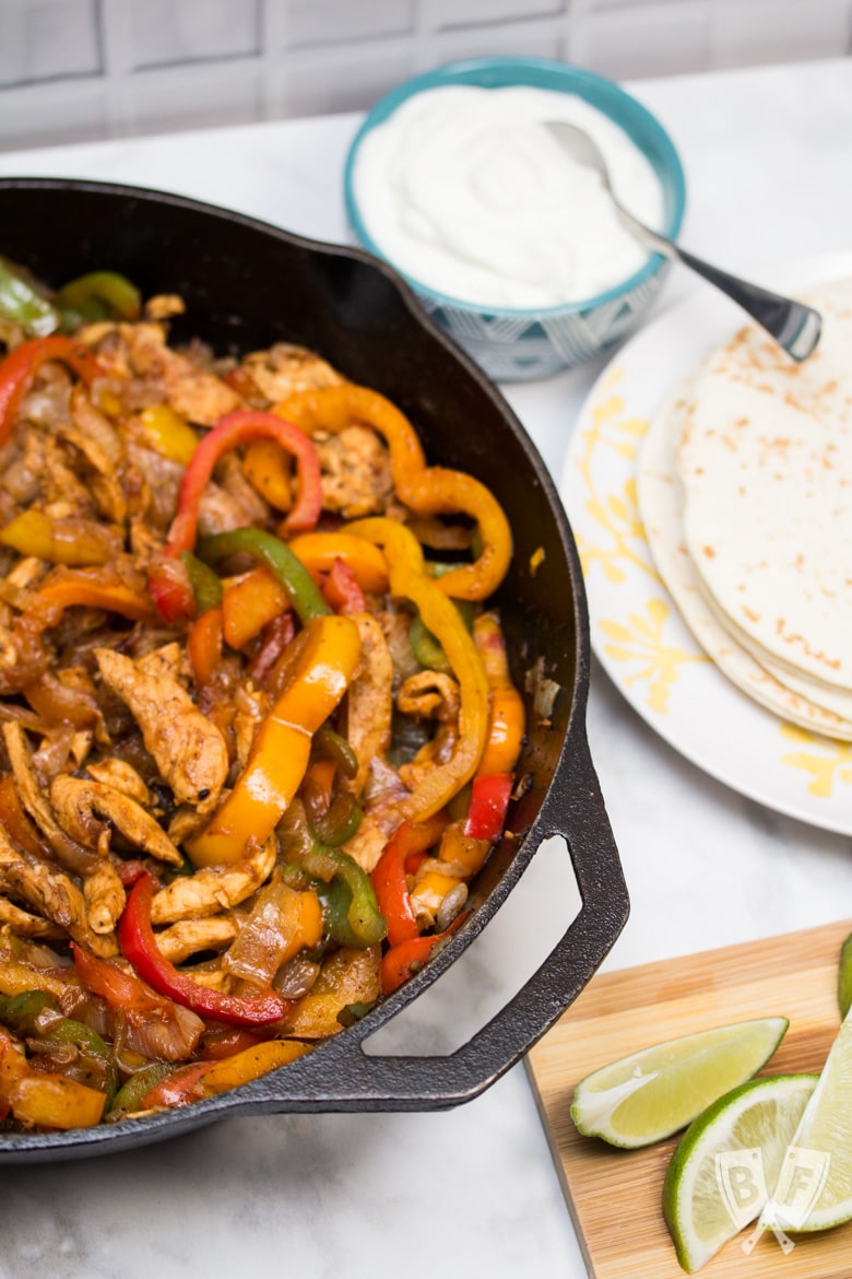 A cast iron skillet filled with chicken fajitas with tortillas and garnishes alongside.