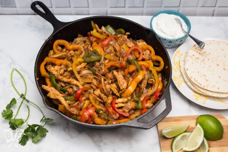 A cast iron skillet filled with chicken fajitas with tortillas and garnishes alongside.