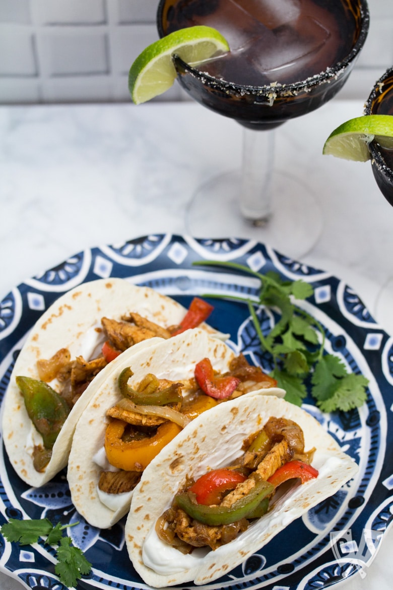 A plate of assembled chicken fajitas with margaritas in the background.