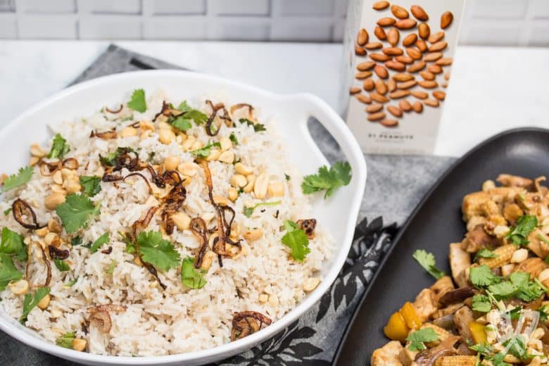 Overhead view of a bowl of Peanutty Rice with Crispy Fried Shallots with a chicken stir-fry to the side and a carton of milked peanuts in the background.