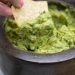 Dipping a chip into freshly made guacamole with margaritas in the background.